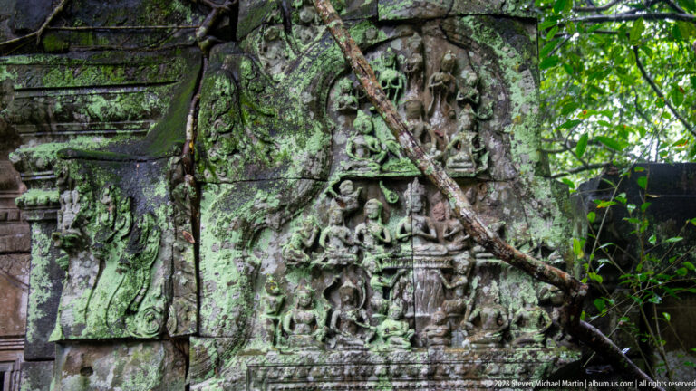 Weathered vertical sandstone panel with stacked carved figures, foliage, and tree roots beside a doorway.