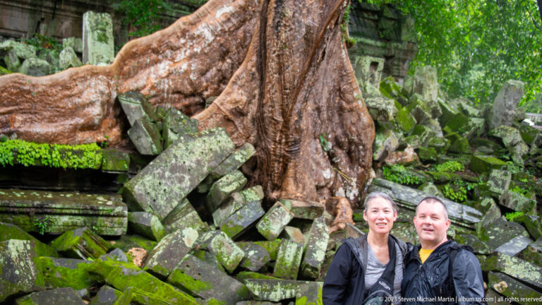 Two visitors stand before a huge tree rooted among mossy collapsed sandstone blocks at Beng Mealea.