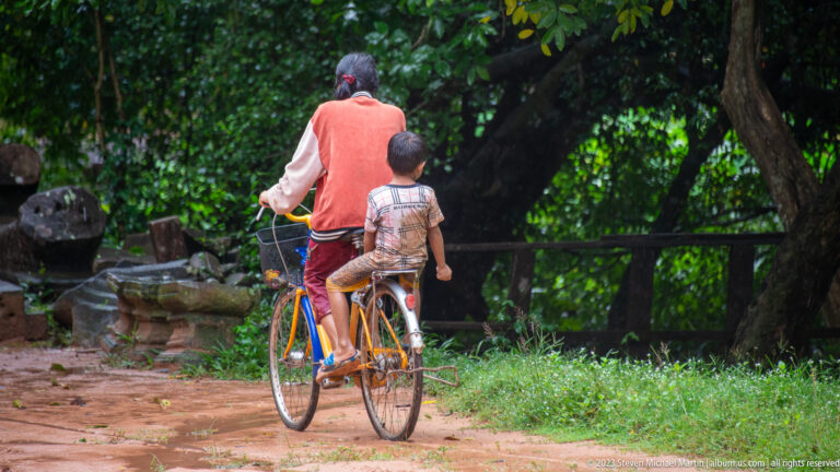 Adult pedals a bicycle along a shaded dirt road with a child seated behind.