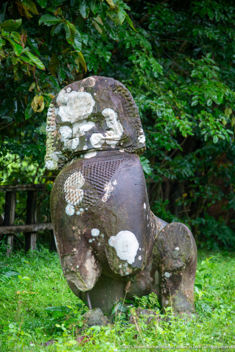 Heavily weathered seated stone guardian animal with pale surface deposits on grass.