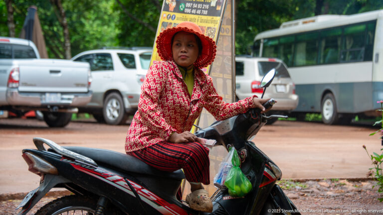 Woman in a red patterned hood sits on a motor scooter beside parked vehicles and signs.