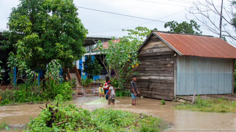 Children stand in floodwater beside timber houses with corrugated-metal walls and roofs.