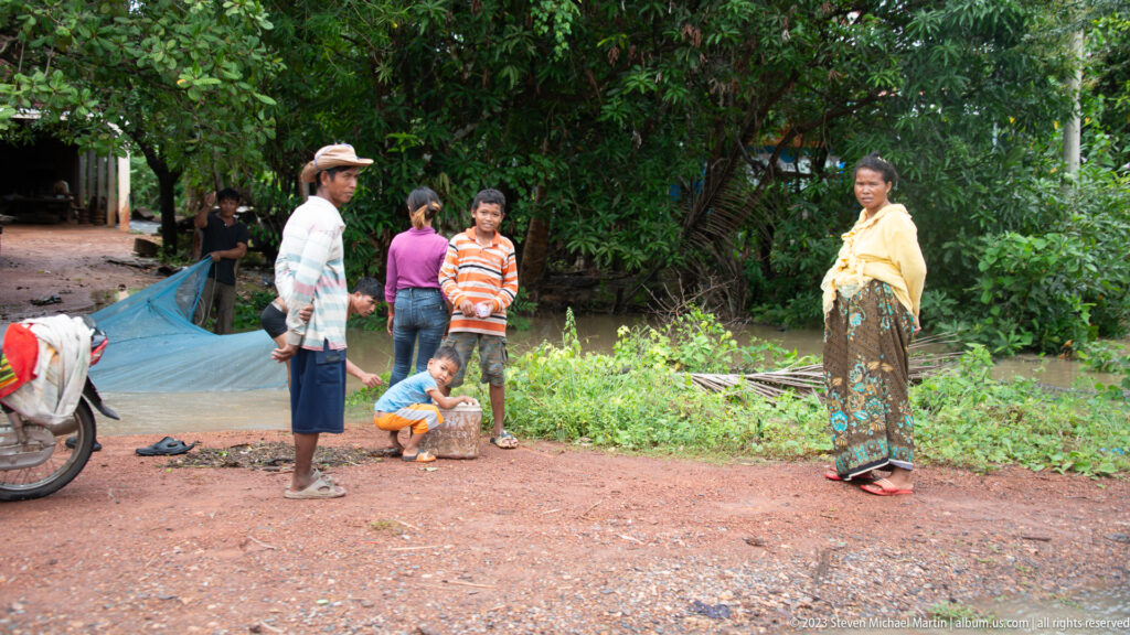 Adults and children gather beside a motorcycle on a shaded red-earth roadside.