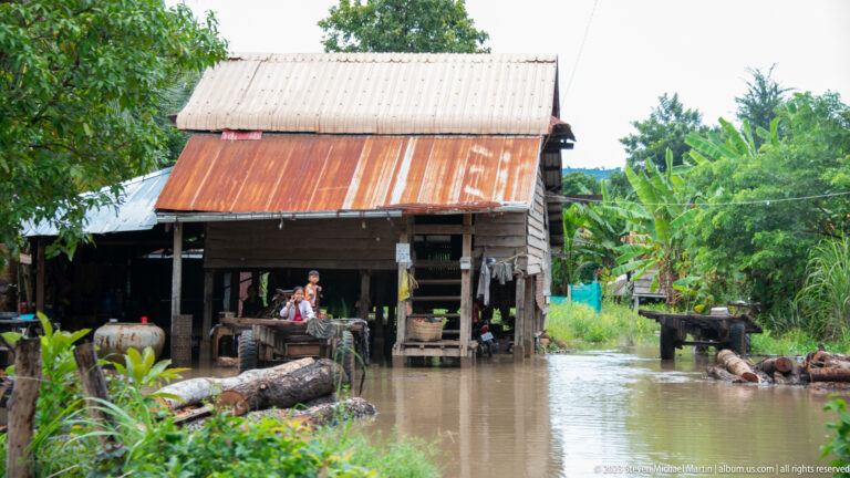 Raised timber house with a corrugated roof stands above floodwater in a rural yard.