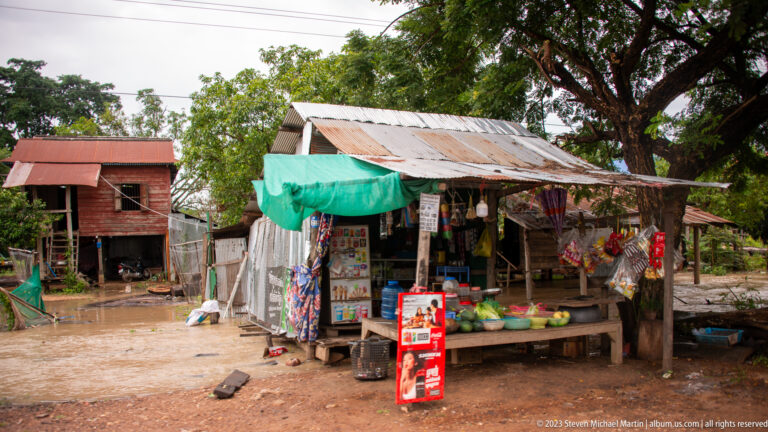 Open-front timber roadside shop displays packaged goods beneath a corrugated roof.