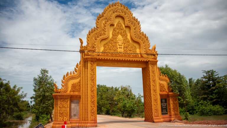 Ornate golden gateway with a tall pointed pediment spans the entrance to a Buddhist compound.