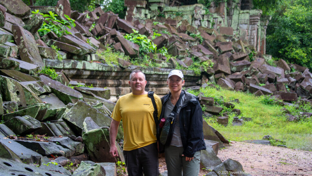 Two visitors standing on wet ground before a large pile of collapsed temple stones.