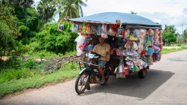 Vendor rides a motorbike covered by a canopy and densely hung colorful household goods.