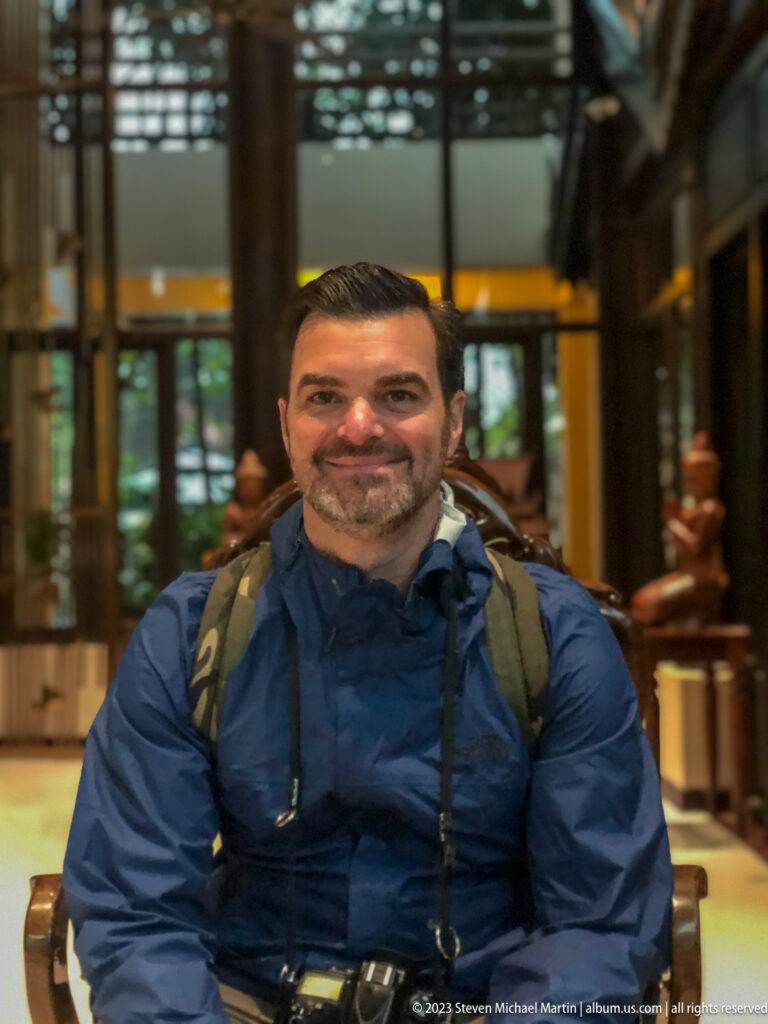 Smiling male traveler in a blue rain jacket and backpack sits before carved figures indoors.