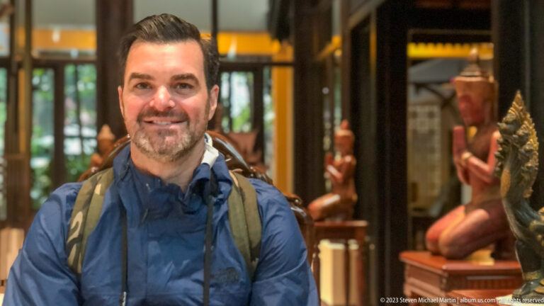 Smiling male traveler in a blue rain jacket and backpack sits before carved figures indoors.
