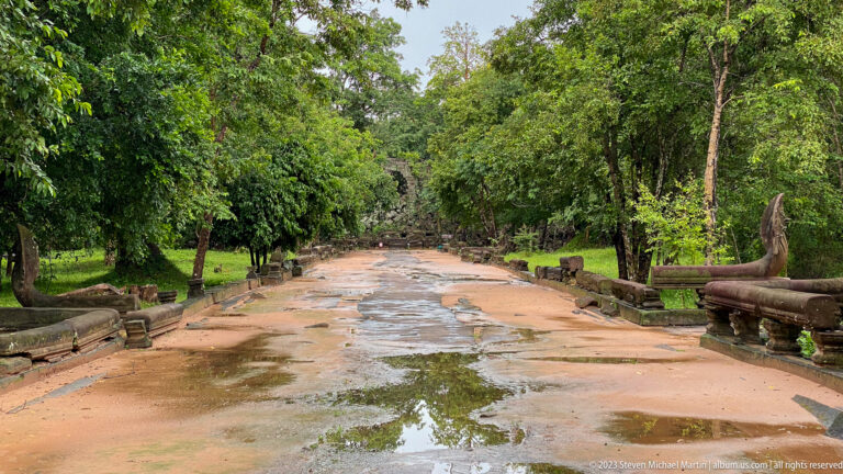 Rain-wet stone approach to Beng Mealea bordered by weathered naga balustrades and dense trees.