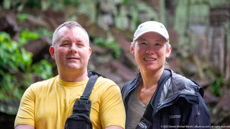Close portrait of two smiling visitors with blurred greenery and temple stone behind them.