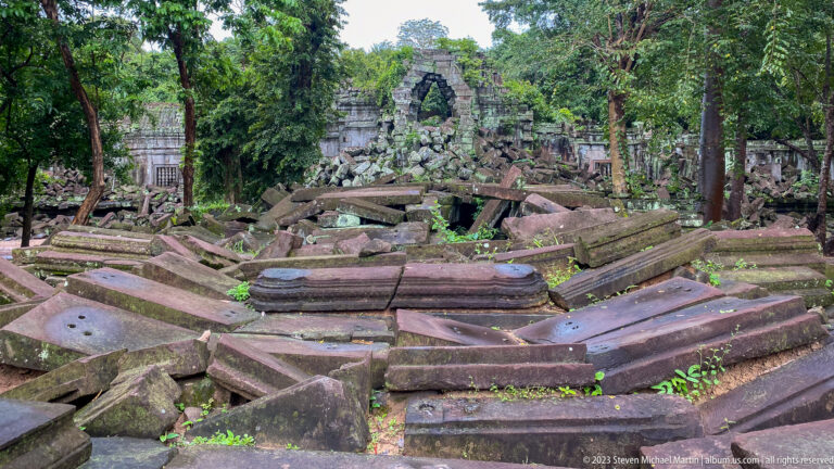 Large field of fallen sandstone blocks and carved architectural fragments before a ruined temple opening.