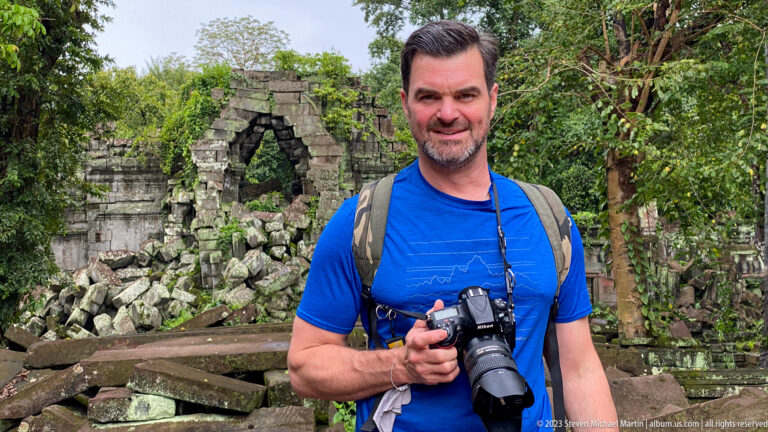 Male traveler holding a camera stands before a collapsed sandstone gateway at Beng Mealea.