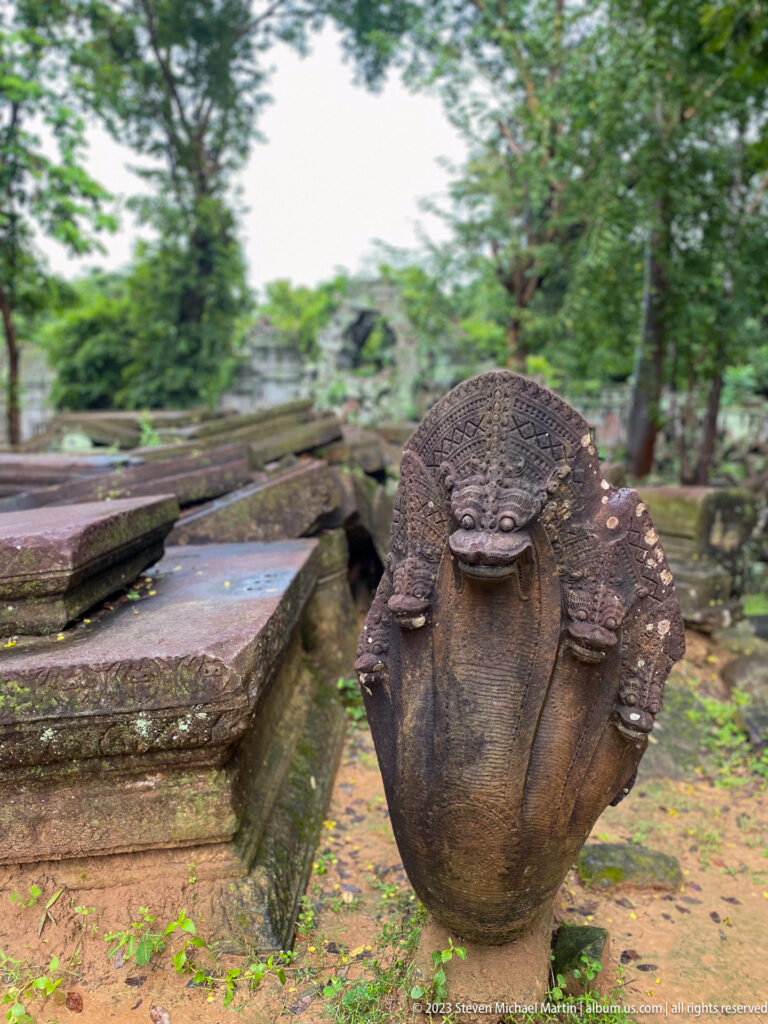 Weathered multi-headed naga stone finial stands beside a ruined temple causeway.