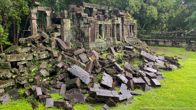 Ruined sandstone gallery with rows of openings rises behind a mound of fallen blocks.