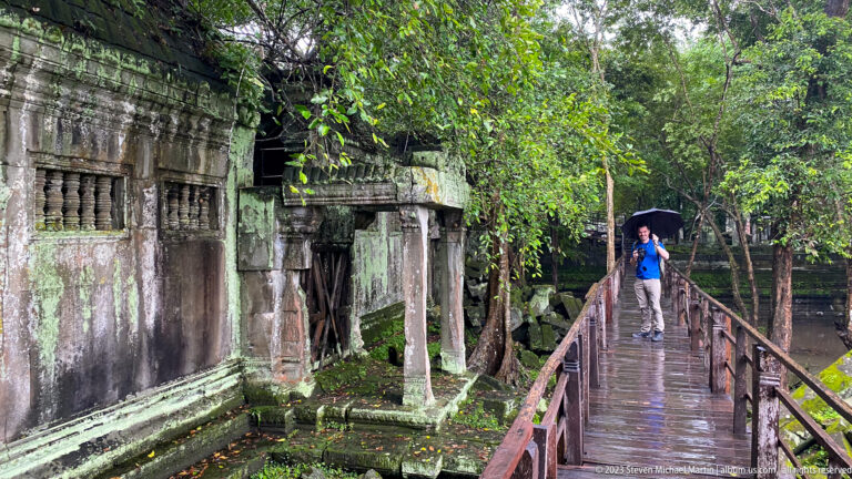 Visitor with an umbrella stands on a wet wooden walkway beside a ruined sandstone gallery.