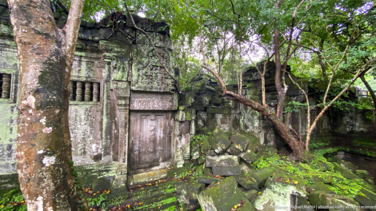 Overgrown sandstone gallery with baluster windows, doorway, fallen blocks, and tree branches.