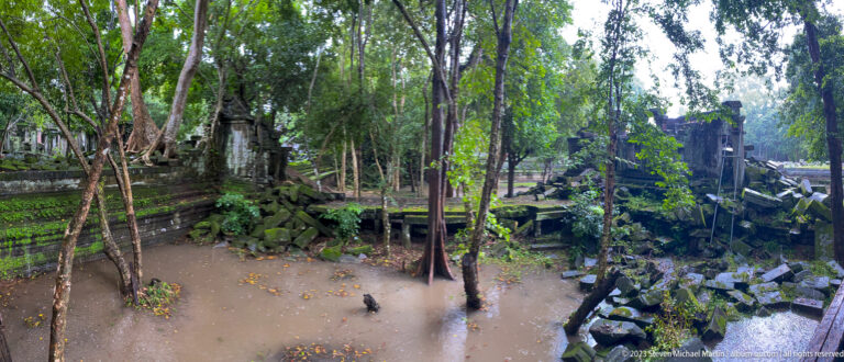 Flooded Beng Mealea courtyard with trees, fallen sandstone, and ruined walls.