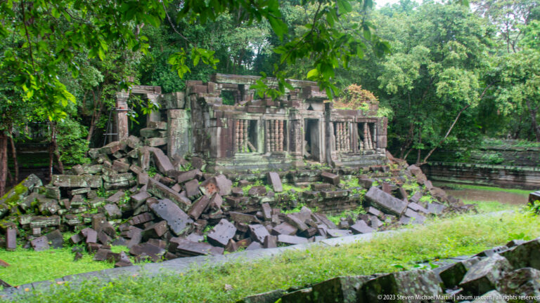 Partly standing rectangular temple building surrounded by piles of fallen sandstone blocks in forest.