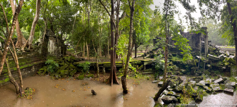 Wide flooded courtyard at Beng Mealea with trees, collapsed stone, and ruined temple walls.
