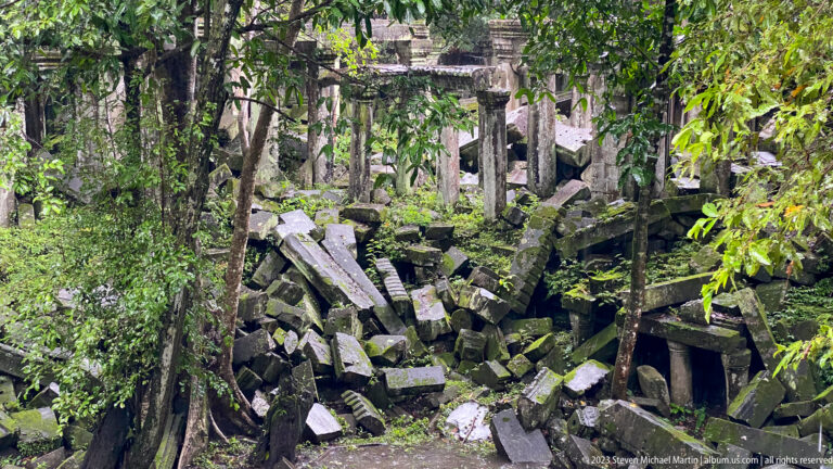 Fallen sandstone blocks fill a ruined gallery seen through trees and dense vegetation.
