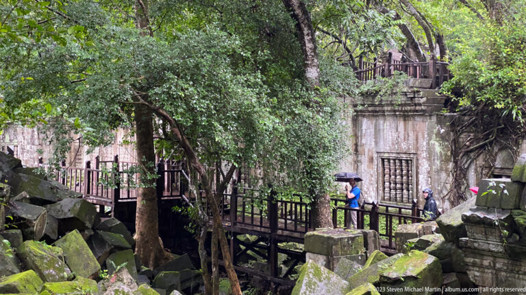 Visitors with umbrellas cross a wooden walkway through collapsed temple ruins and trees.