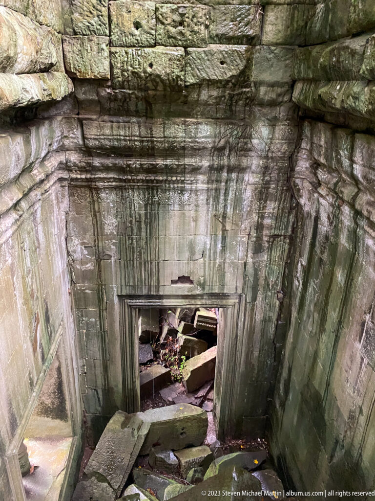Sunken sandstone chamber descends toward a low doorway blocked by fallen stones.