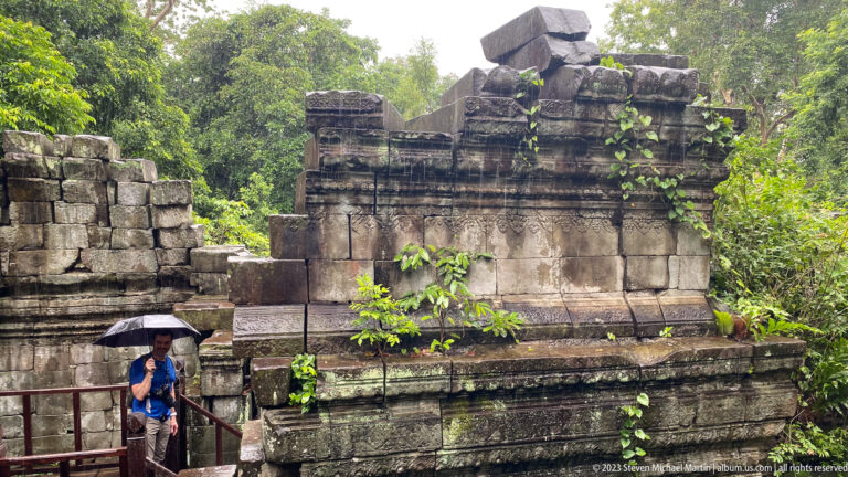 Compact sandstone temple structure with a molded cornice rises beside a visitor holding an umbrella.