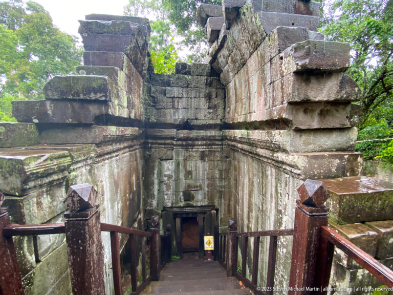 Wooden stairs descend between collapsed sandstone walls into a lower section of Beng Mealea.