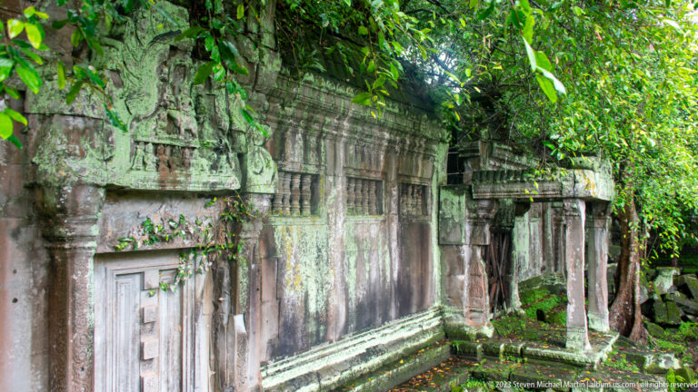 Oblique view along a moss-streaked sandstone gallery wall with pilasters, doorways, and a small porch.