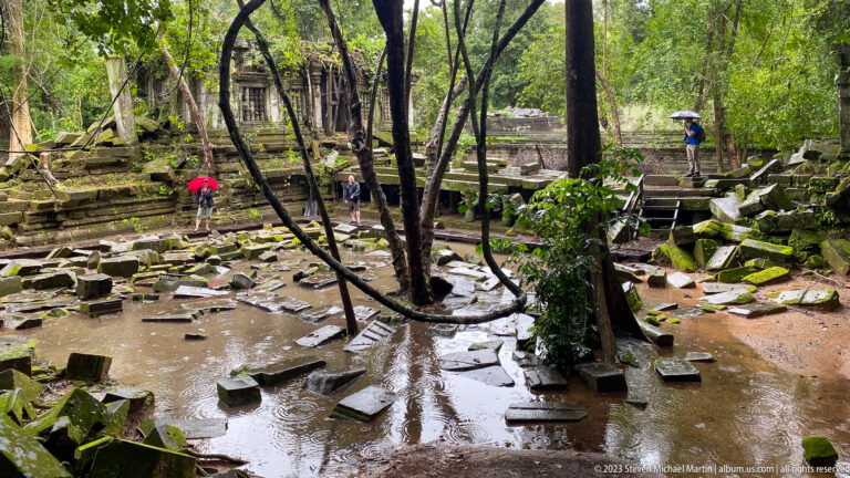 Visitors cross raised walkways above a flooded courtyard scattered with sandstone blocks and trees.