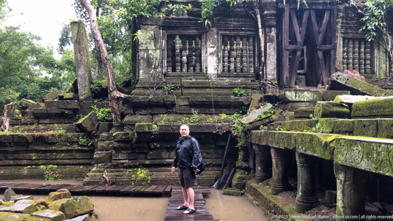 Male traveler stands on a narrow walkway above floodwater before a ruined gallery with stone-baluster windows.