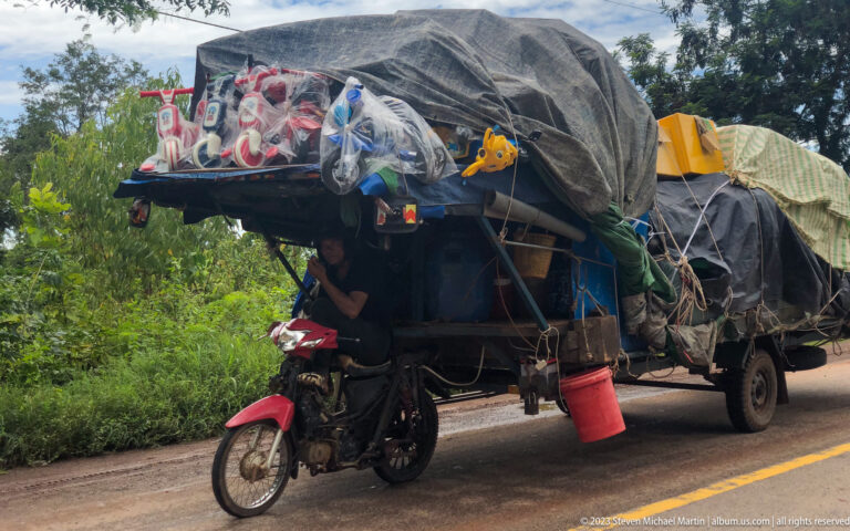 Motorcycle-powered cargo vehicle carries a large tarpaulin-covered load and household goods along a road.