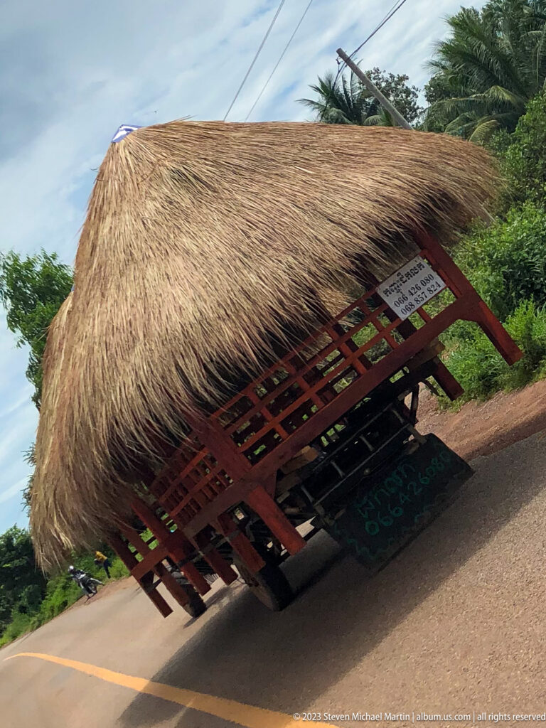 Large palm-thatch roof structure with a red frame is transported along a rural Cambodian road.