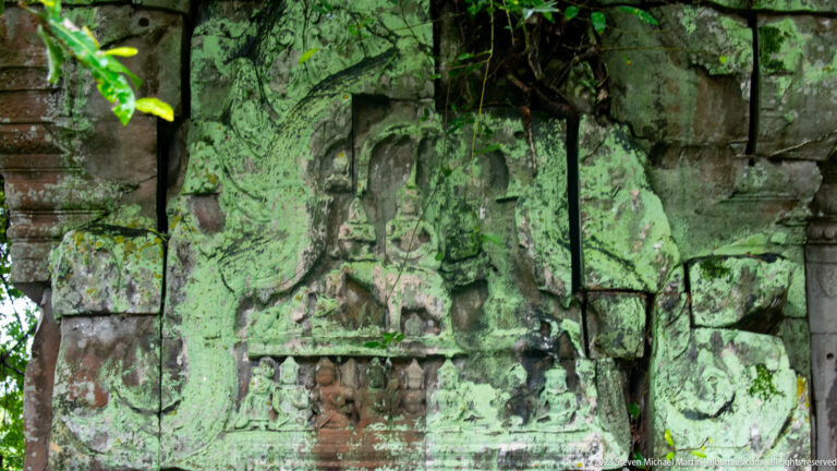Weathered standing female figure carved on green lichen-covered sandstone blocks within the temple wall.