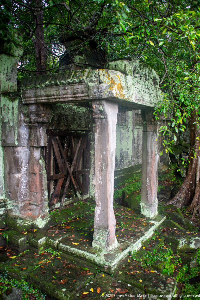 Low mossy sandstone porch with four posts and timber bracing beneath dense foliage.