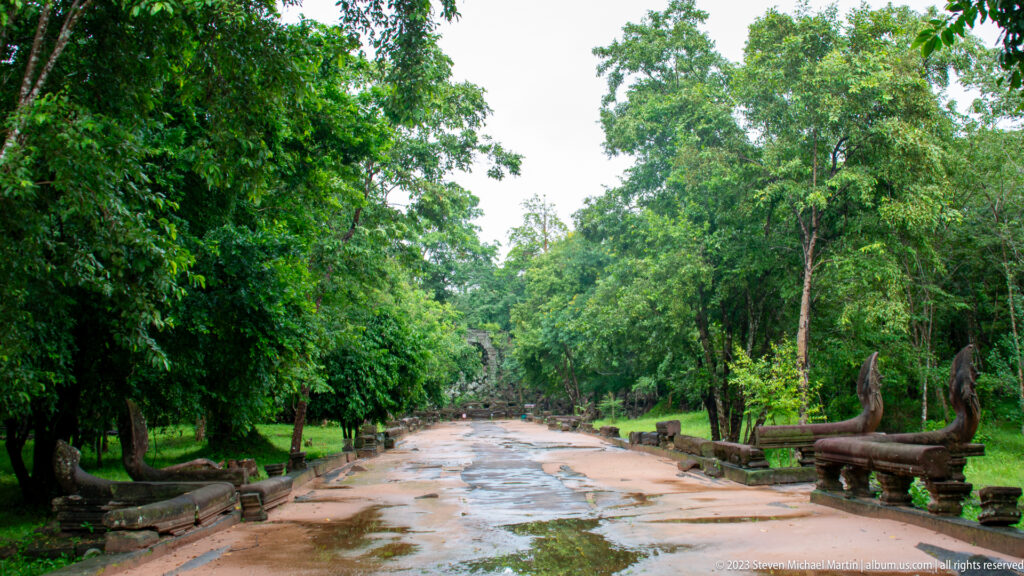Wet stone causeway through dense forest with surviving naga balustrade sections along both sides.