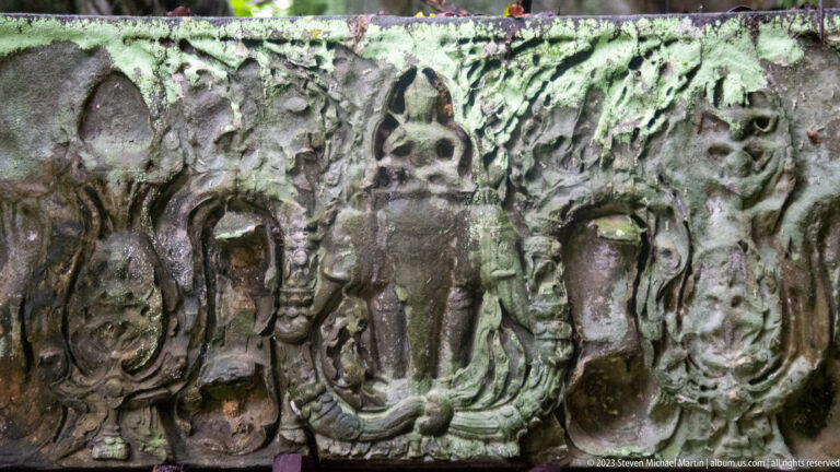 Weathered sandstone relief crowded with several carved figures arranged around a central vertical form.