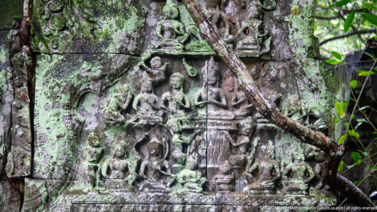 Vertical sandstone relief with one central standing figure and several smaller figures arranged around it.