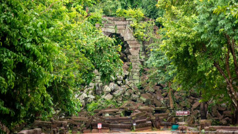 Partly standing stone gateway behind a dense pile of collapsed sandstone blocks in tropical vegetation.