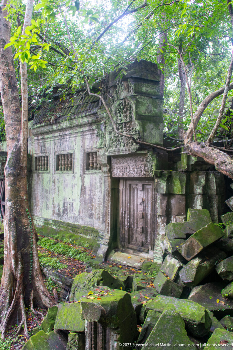 Mossy sandstone façade with a false door, baluster windows, tree roots, and collapsed blocks.