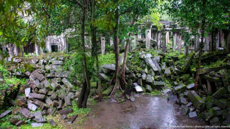 Wide view of trees and collapsed sandstone filling a court between surviving temple galleries.
