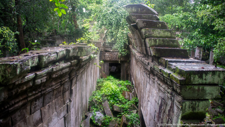 Narrow corridor between sandstone walls filled with fallen roof stones, moss, and vegetation.