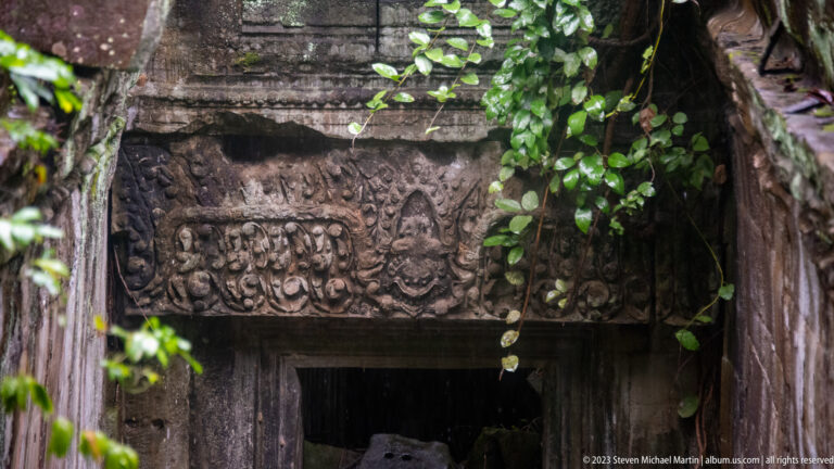 Carved sandstone lintel with a central mask and symmetrical foliage above a dark doorway.
