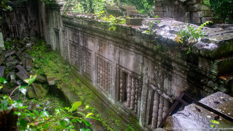 Long sandstone gallery wall with repeated baluster windows, pilasters, moss, and fallen blocks.