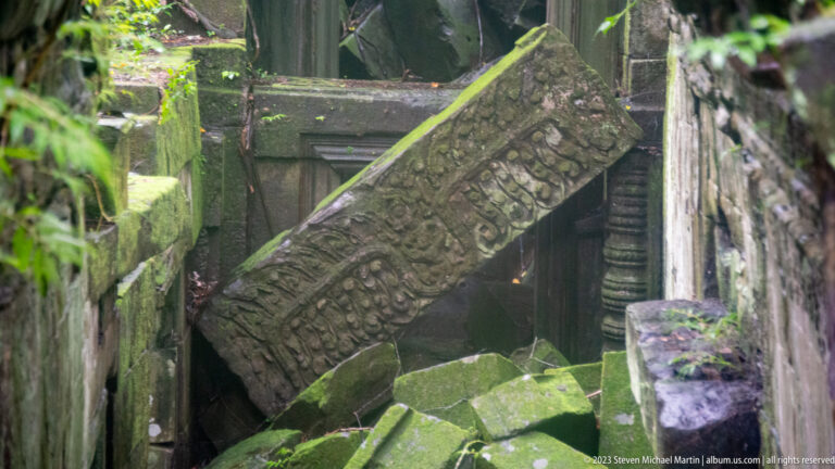 Carved sandstone lintel fallen diagonally across a blocked passage amid mossy temple rubble.