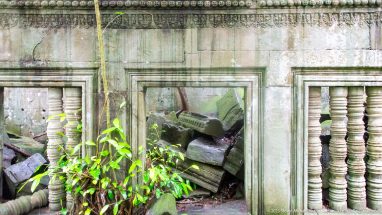 Sandstone wall with two baluster windows and a central doorway blocked by fallen stones.