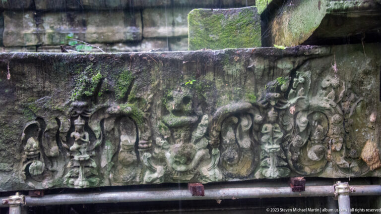 Weathered carved lintel with seated figures, scrolling foliage, and a central mask-like motif.