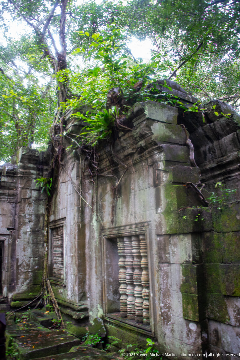 Oblique view of a sandstone gallery corner with baluster window and vegetation-covered roof.